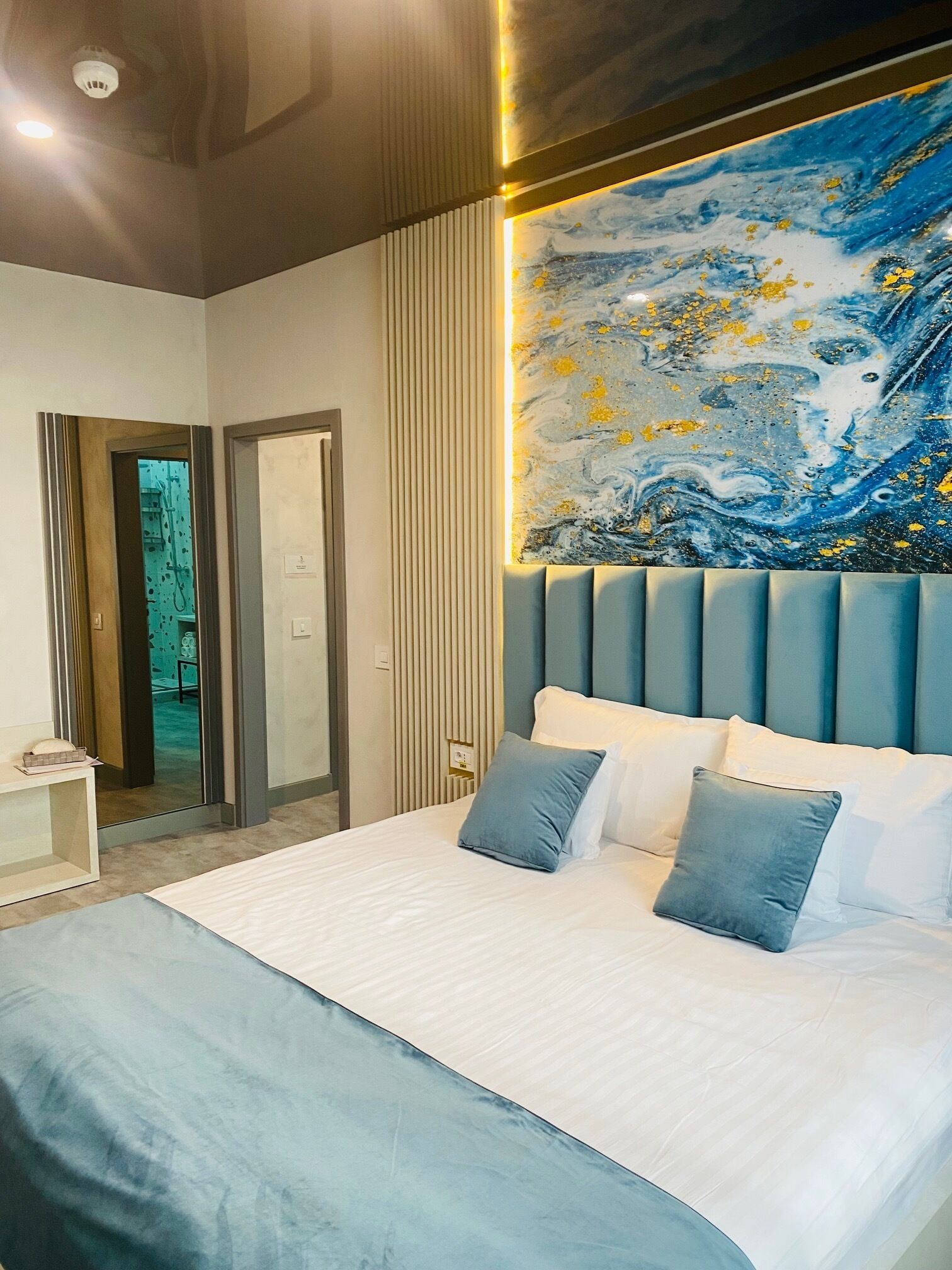 Una habitación de hotel moderna con una cama grande, un cabecero con panel azul, una obra de arte colgada en la pared y un baño visible a través de una puerta.