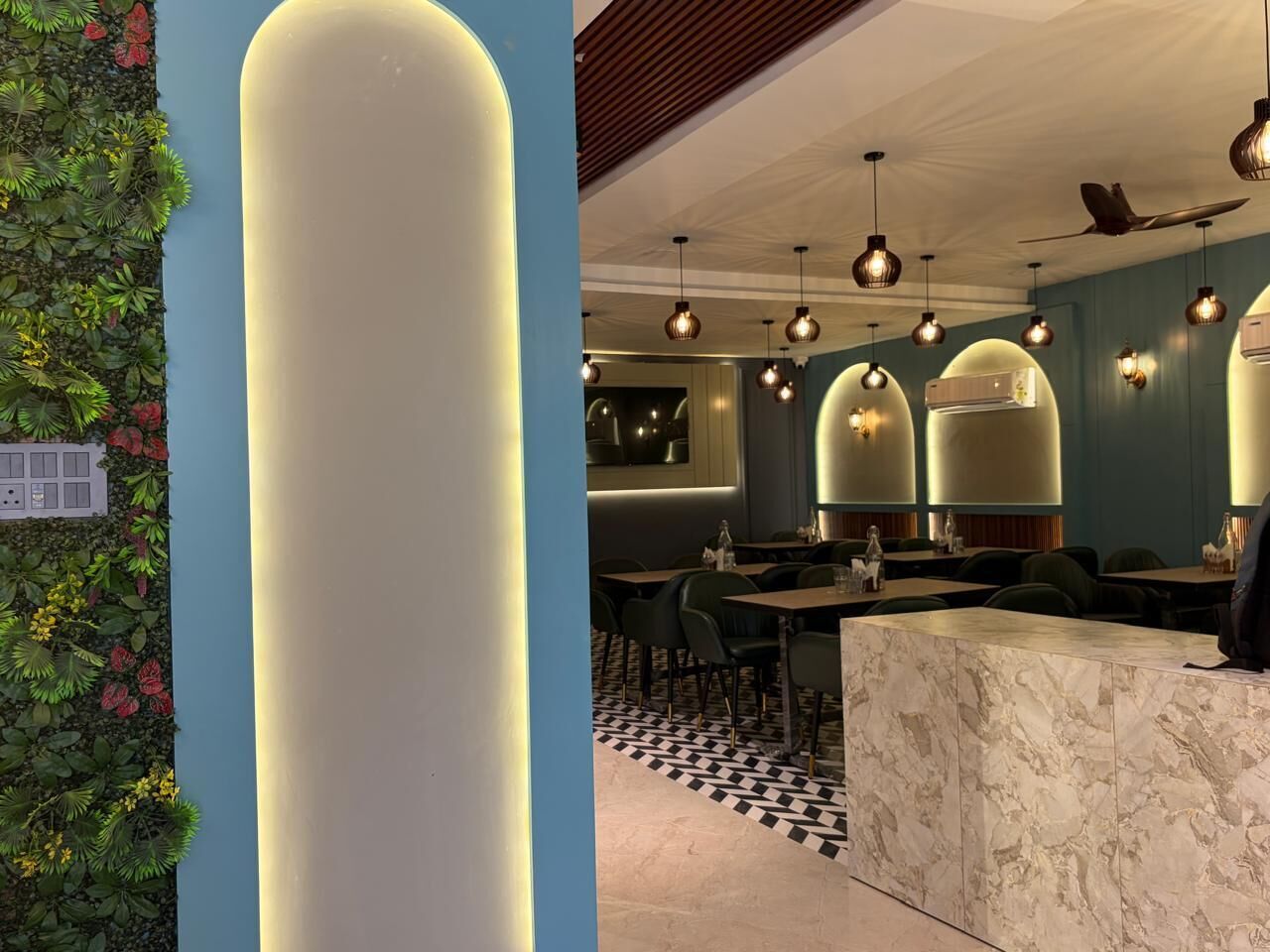 A restaurant interior with a marble reception desk, green chairs, and pendant lighting.
