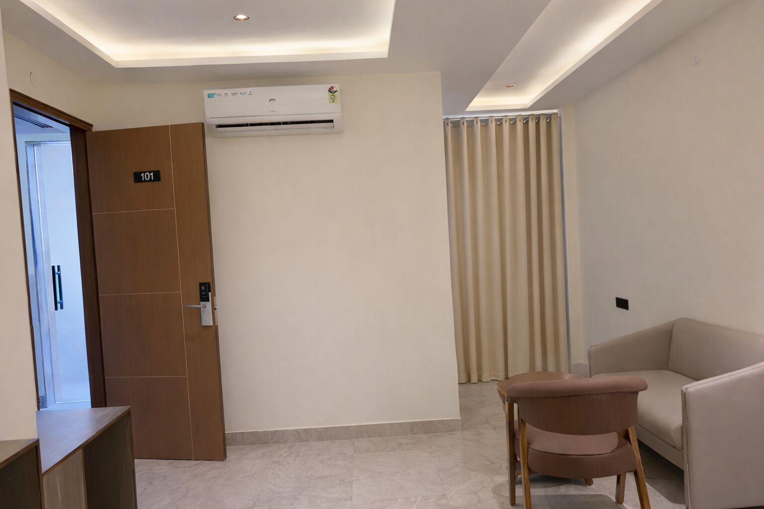 A hotel room with a door labeled '101', an air conditioning unit, a beige curtain, and a beige sofa.