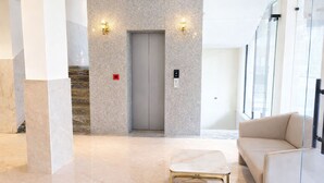 A modern lobby with a marble floor, a beige sofa, a gold-framed table, and a glass elevator. - The Prominence Premium Hotel & Banquet  (Greater Noida)