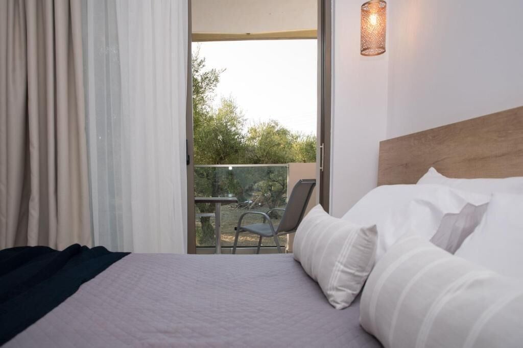 A hotel room with a bed, a chair, a balcony with a view of trees, and a wall-mounted light fixture.