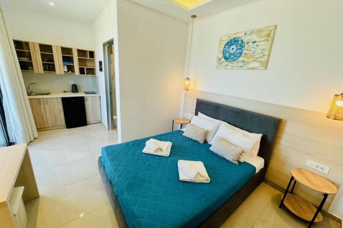 A modern hotel room with a large bed, a small kitchen area, and a painting on the wall.