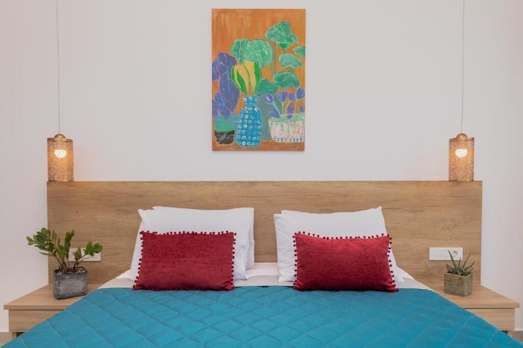 A neatly made bed with a blue quilt, two red pillows, and a wooden headboard. There are two small bedside tables with potted plants. A colorful painting hangs above the bed.
