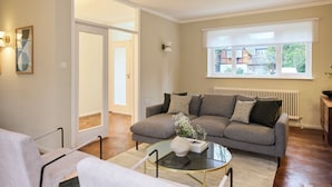 Een woonkamer met een grijze bank, een glazen salontafel en een witte fauteuil. Er is een raam met uitzicht op huizen.