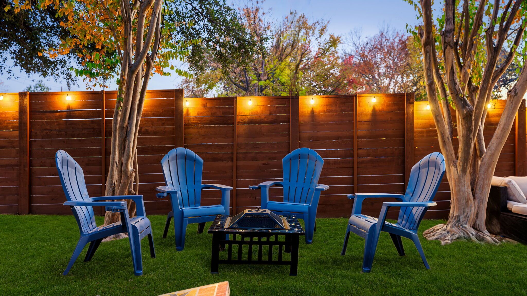 A backyard with a wooden fence, four blue chairs, a small table, and a tree.