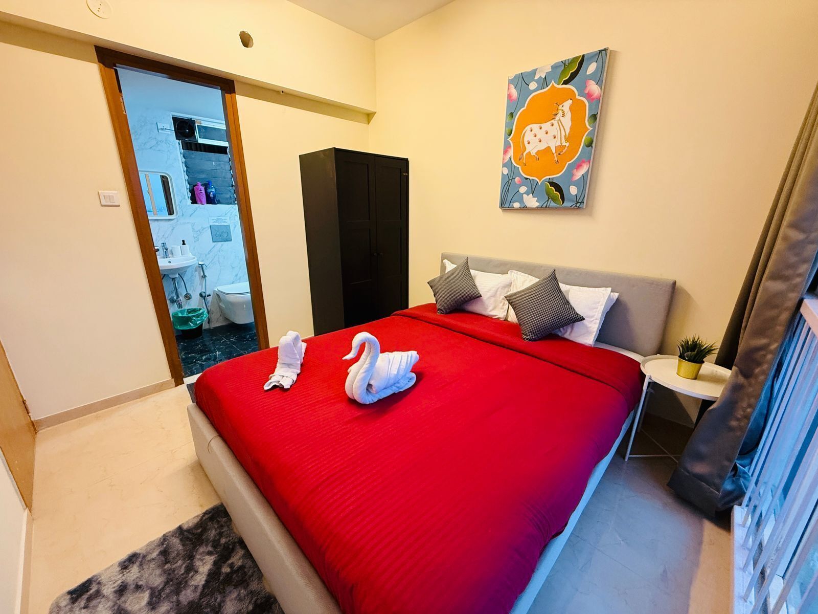 Une chambre d’hôtel avec une couvre-lit rouge, deux serviettes, une tête de lit grise, une petite table de chevet avec une plante et un tableau au mur.