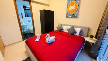 A hotel room with a red bedspread, two towels, a grey headboard, a small bedside table with a plant, and a painting on the wall.