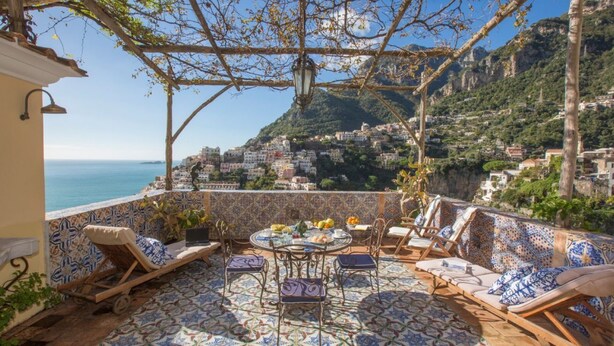 A terrace with a view of the sea and a mountainous town, furnished with lounge chairs and a table. - Residenza Crocione - Terrace sea view (Positano)