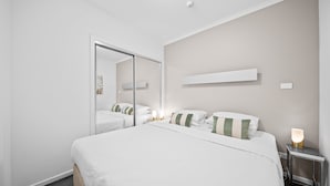 A modern hotel room with a large bed, a bedside table with a lamp, a mirror, and a wall-mounted shelf. - Bright 1-bed Apartment in Greenway (Canberra)