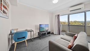 A modern living room with a grey sofa, a blue chair, a TV on a stand, and a large window with a view of trees and hills. - Bright 1-bed Apartment in Greenway (Canberra)