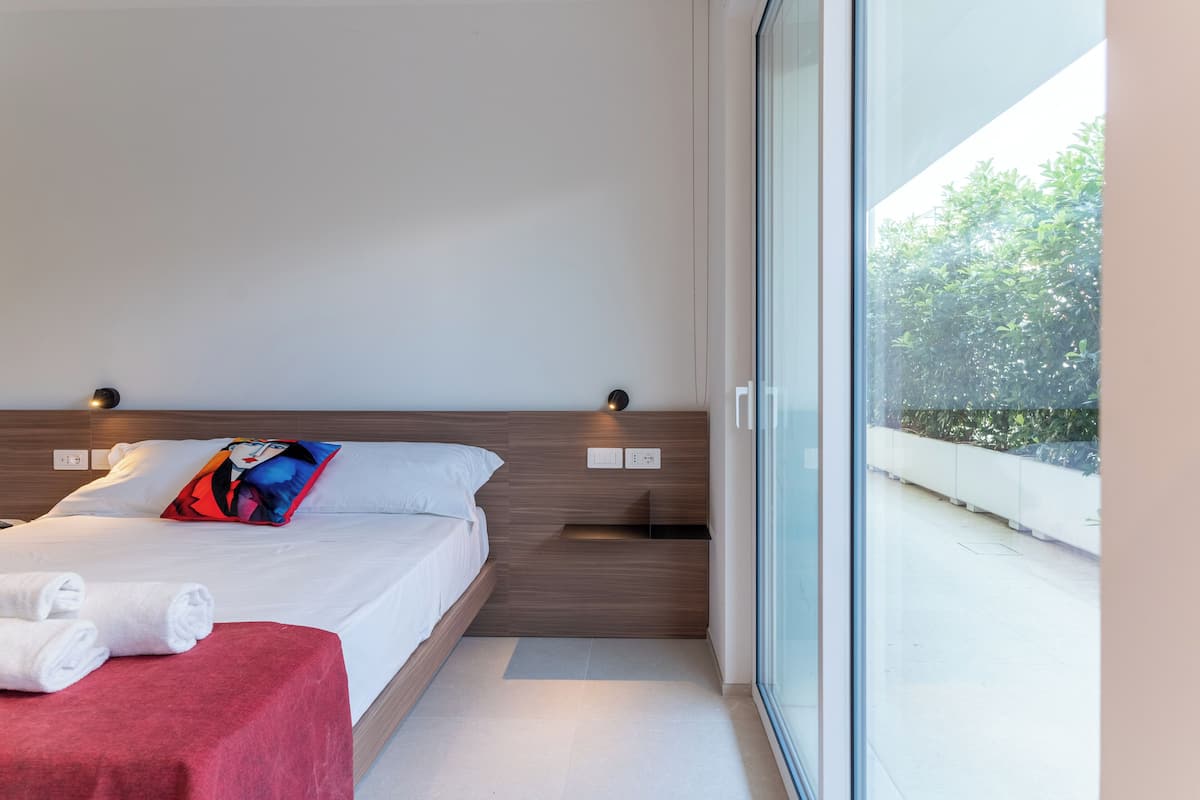 A modern hotel room with a large bed, a red accent bed runner, a pillow with a graphic design, and a view of greenery through a glass door.