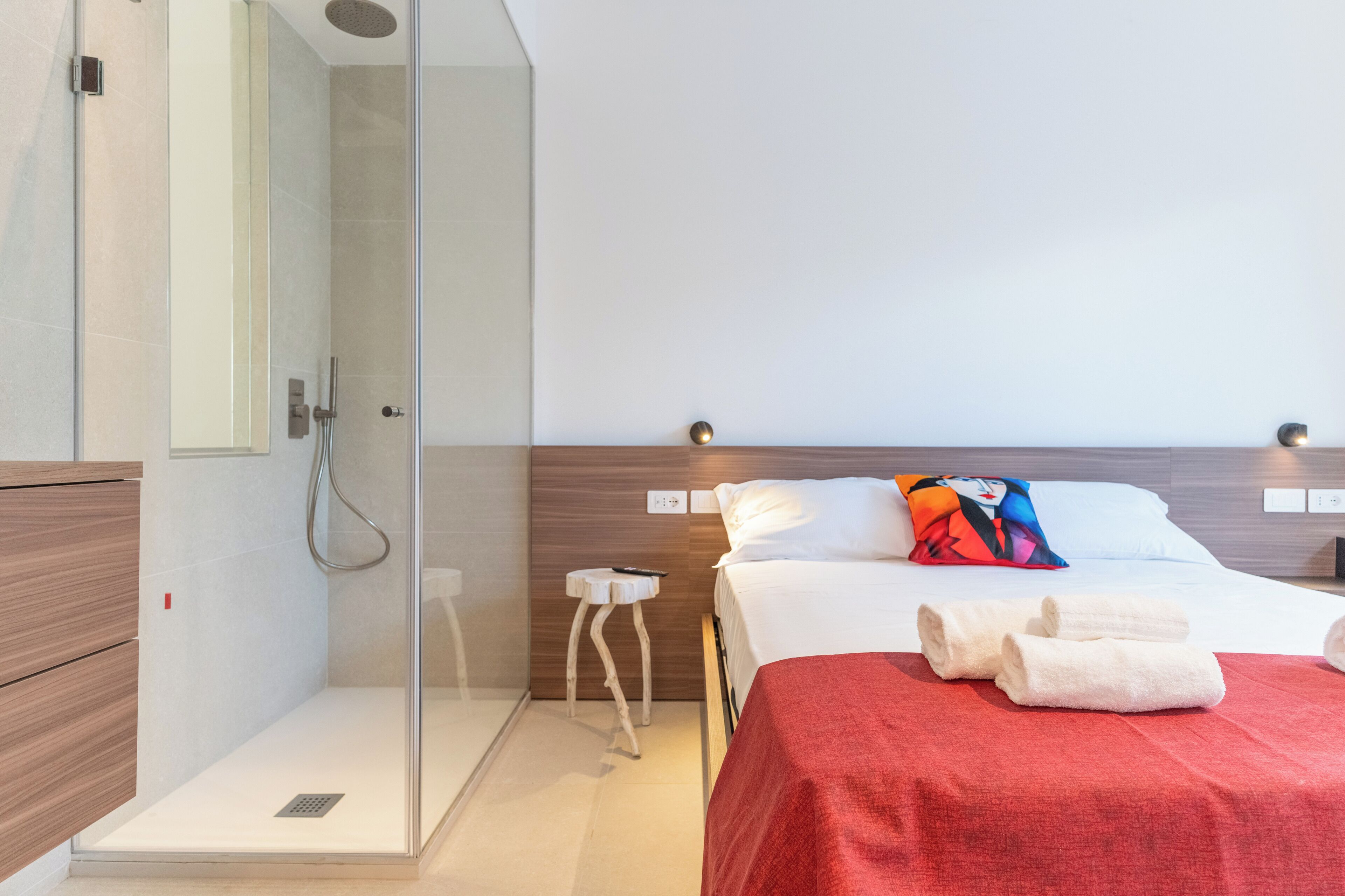 Habitació d'hotel moderna amb dutxa envidriada, armari de fusta, taula de nit amb comandament a distància, llit amb roba blanca i coixí de colors, i dues tovalloles arremolinades al llit.