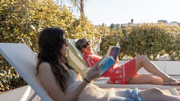 Two individuals reclining on lounge chairs, reading books, with a hedge and a building in the background.