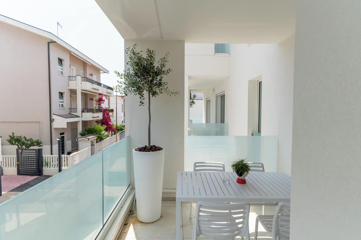 A balcony with a white table and chairs, a potted plant, and a view of neighboring buildings.