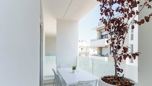 Un balcon avec une table et des chaises blanches, un pot de fleurs et une vue sur les bâtiments voisins.