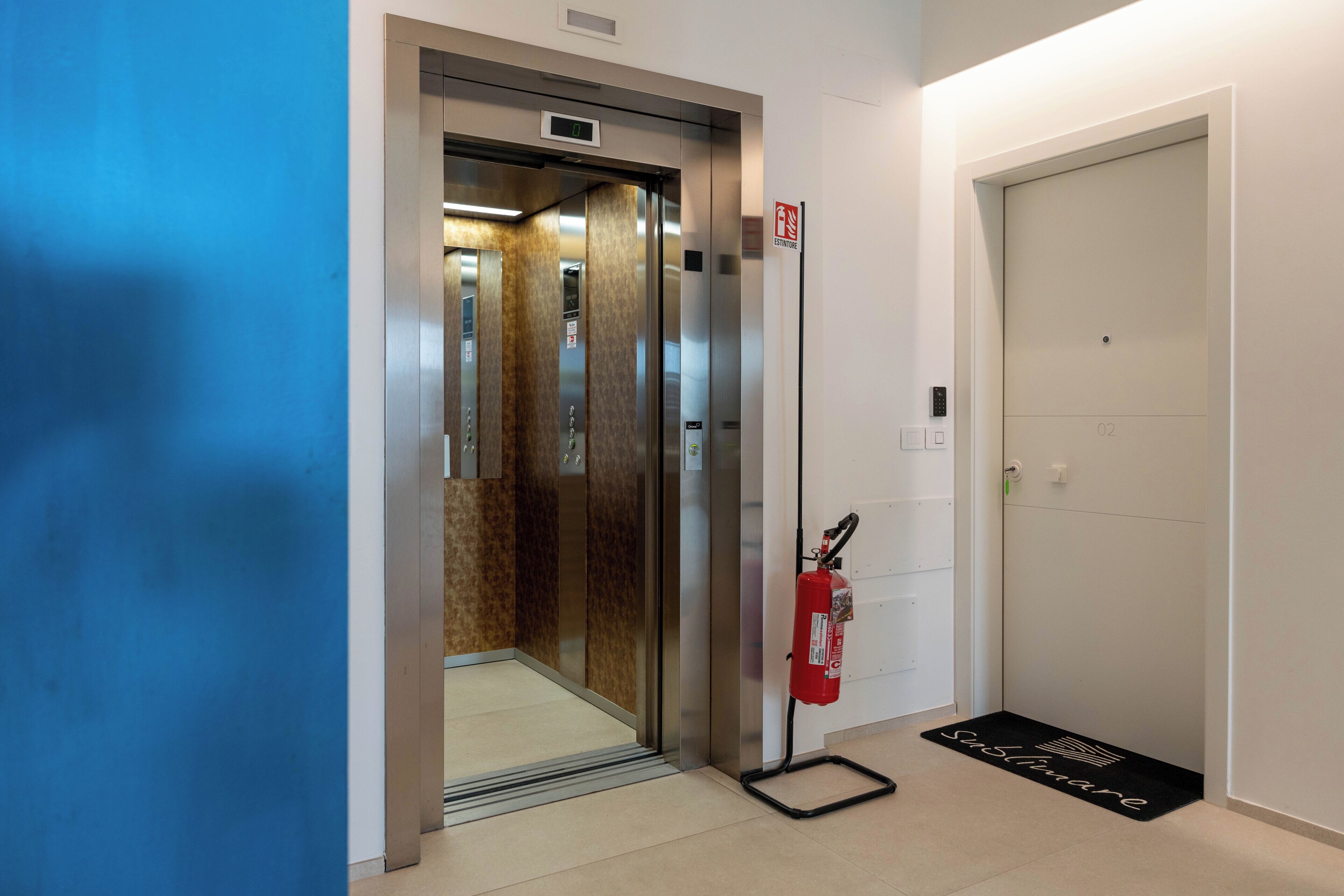 An elevator with a fire extinguisher mounted on the wall, a doormat with the hotel's name, and a door with a number indicating the floor.