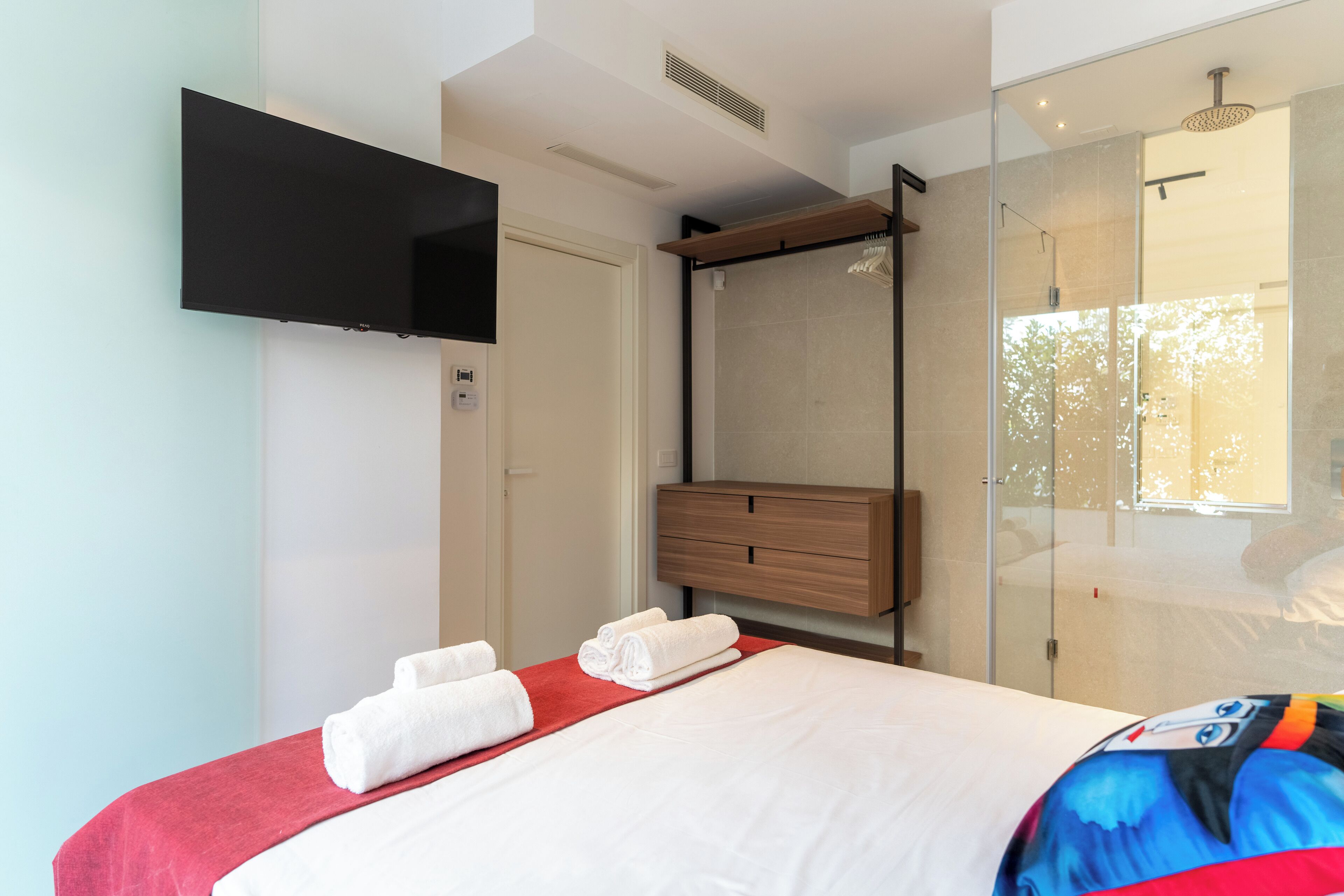 A modern hotel room with a bed, a mounted TV, a shower area, and a wooden dresser.