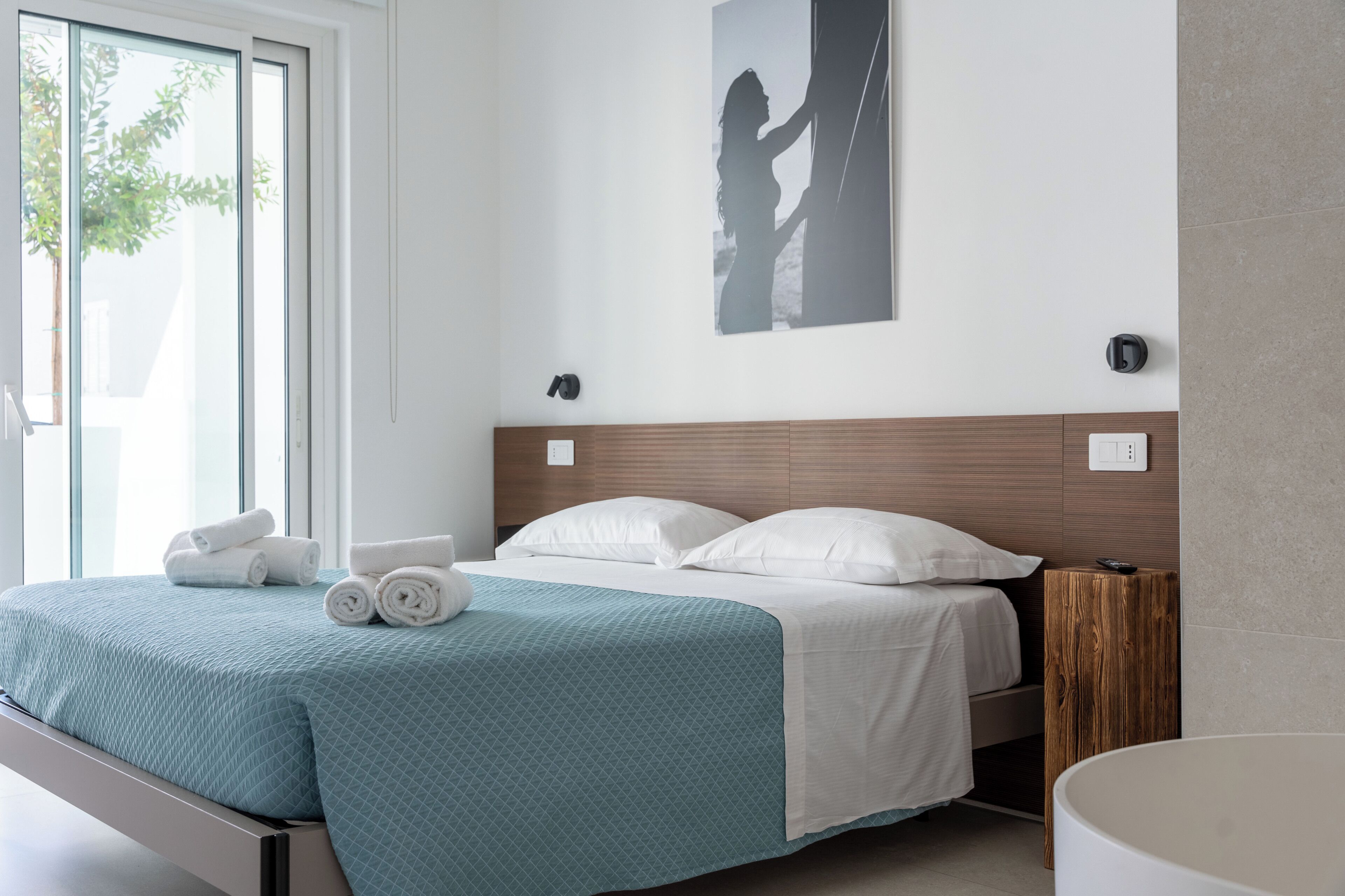 Habitació d'hotel amb un llit, una làmpada penjada a la paret i una finestra amb vista a arbres.
