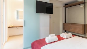 Une chambre d’hôtel avec un lit, un téléviseur fixé au mur, une commode en bois et une salle de bain visible à travers une porte ouverte.