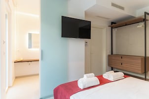 Habitación de hotel con cama, televisión montada en la pared, cómoda de madera y baño visible a través de una puerta abierta.