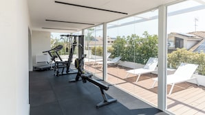 Une salle de sport moderne, équipée de matériel d’exercice, avec vue sur un patio agrémenté de chaises longues blanches.