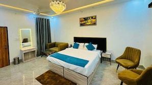 A modern bedroom with a large bed, a chandelier, a ceiling fan, a nightstand, a chair, a desk, and a mirror. - THE GRAND PALACE (Prayagraj)