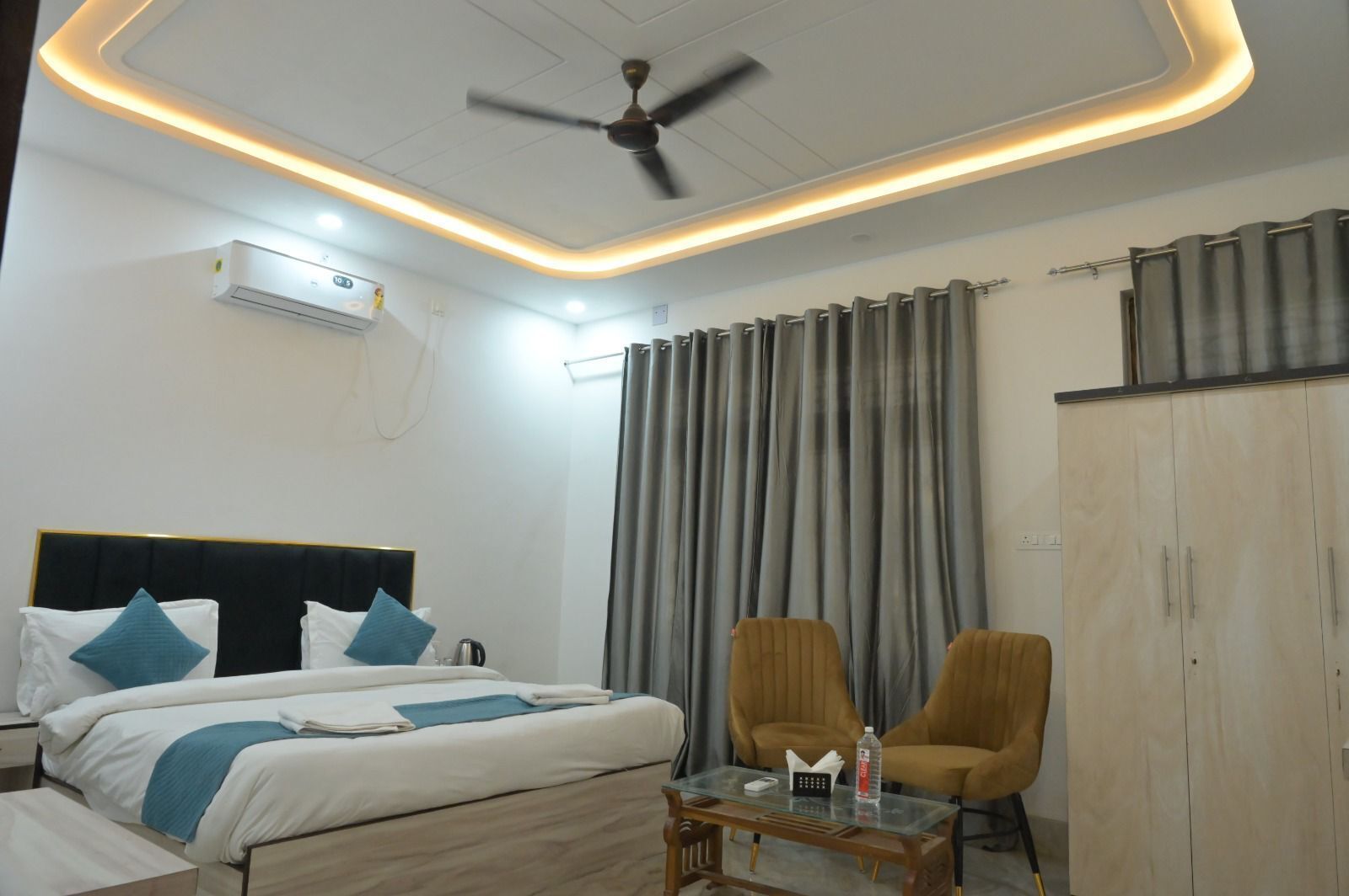 A hotel room with a bed, a ceiling fan, a table with a bottle, two chairs, and a wardrobe.