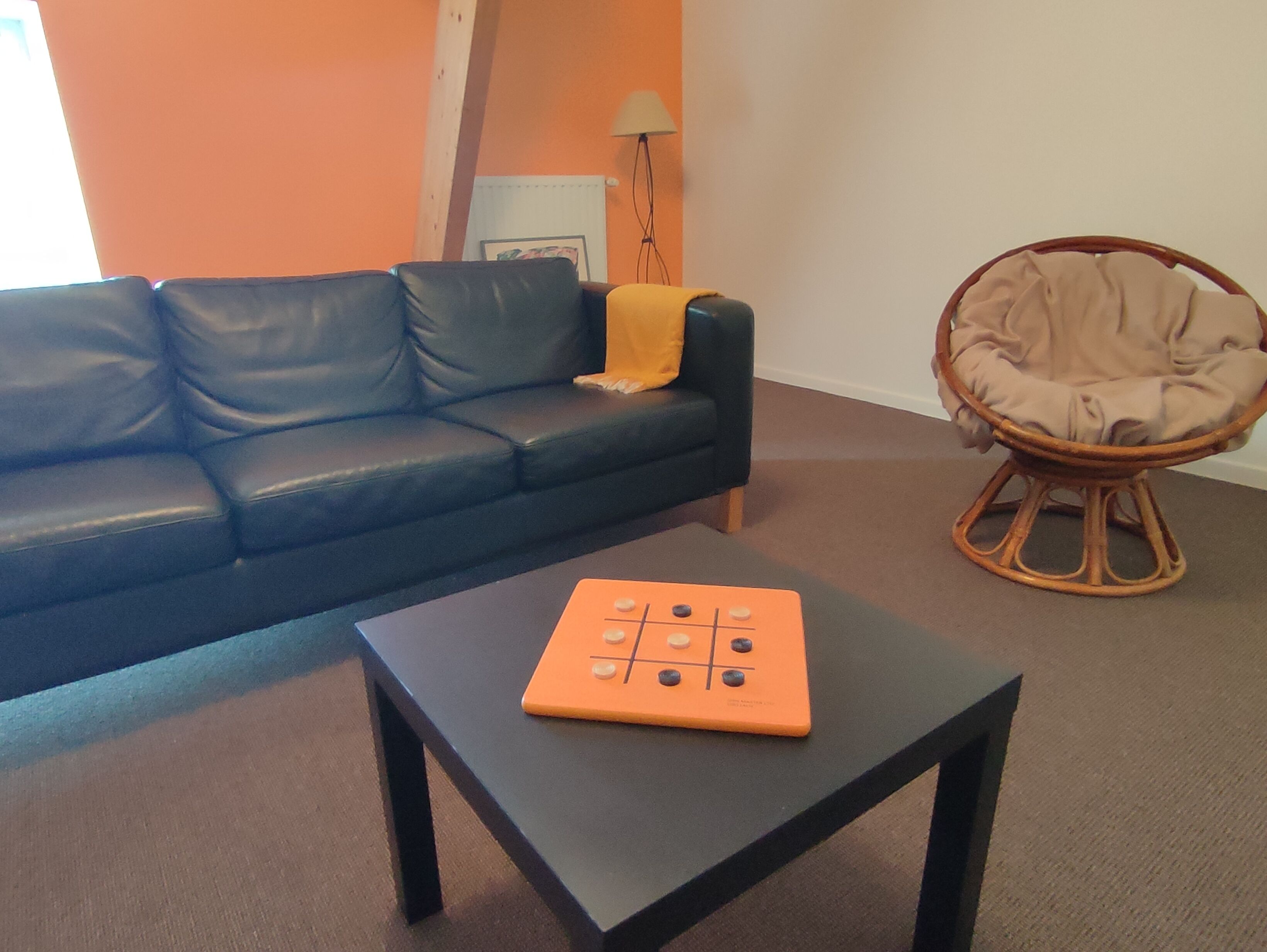 Ein Wohnzimmer mit einem schwarzen Ledersofa, einem kleinen schwarzen Couchtisch mit einem Tic-Tac-Toe-Spiel, einem Korbstuhl und einer orangefarbenen Wand.
