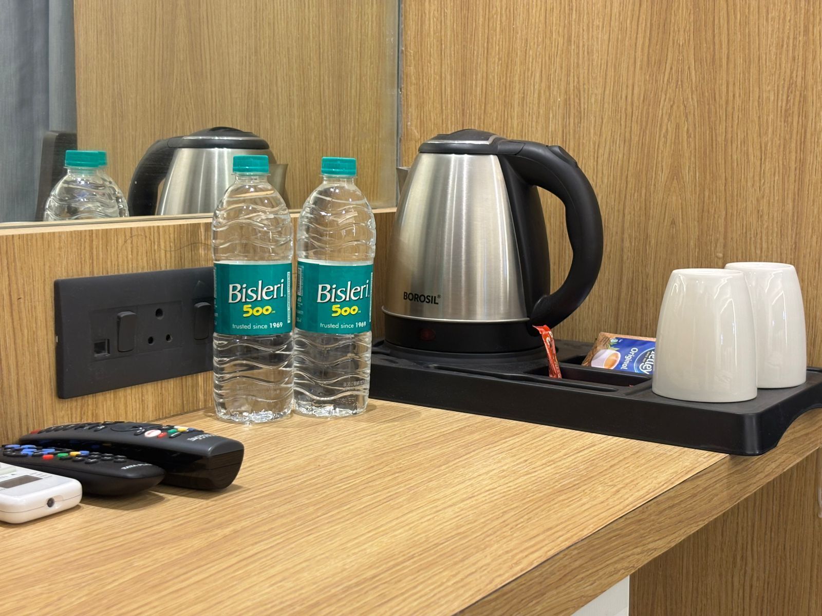 A hotel room counter with a coffee maker, two Bisleri water bottles, a remote control, a white mug, and a packet of tissues.