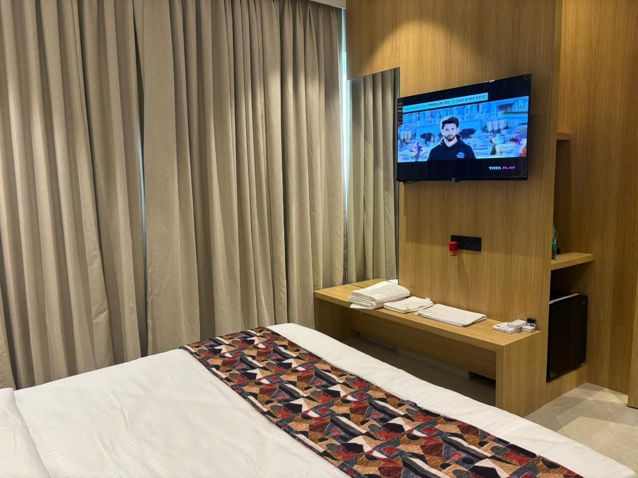 A hotel room with a bed, a TV mounted on the wall, a wooden bench with towels, and a curtain.