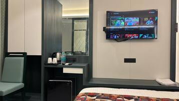 A hotel room with a bed, a desk with a computer, a chair, a mirror, and a TV mounted on the wall.