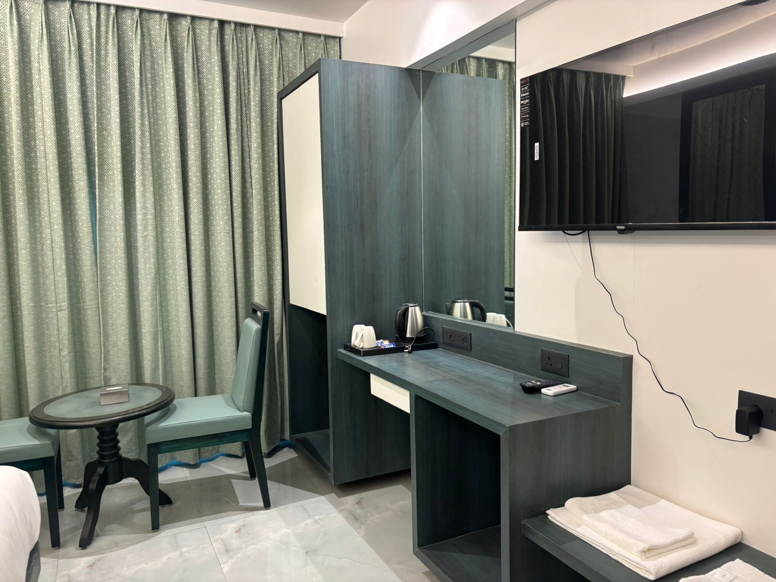 Ein Hotelzimmer mit Bett, einem kleinen runden Tisch, einem Stuhl, einem Schreibtisch mit Spiegel, einem an der Wand montierten Fernseher und einem Handtuchhalter.