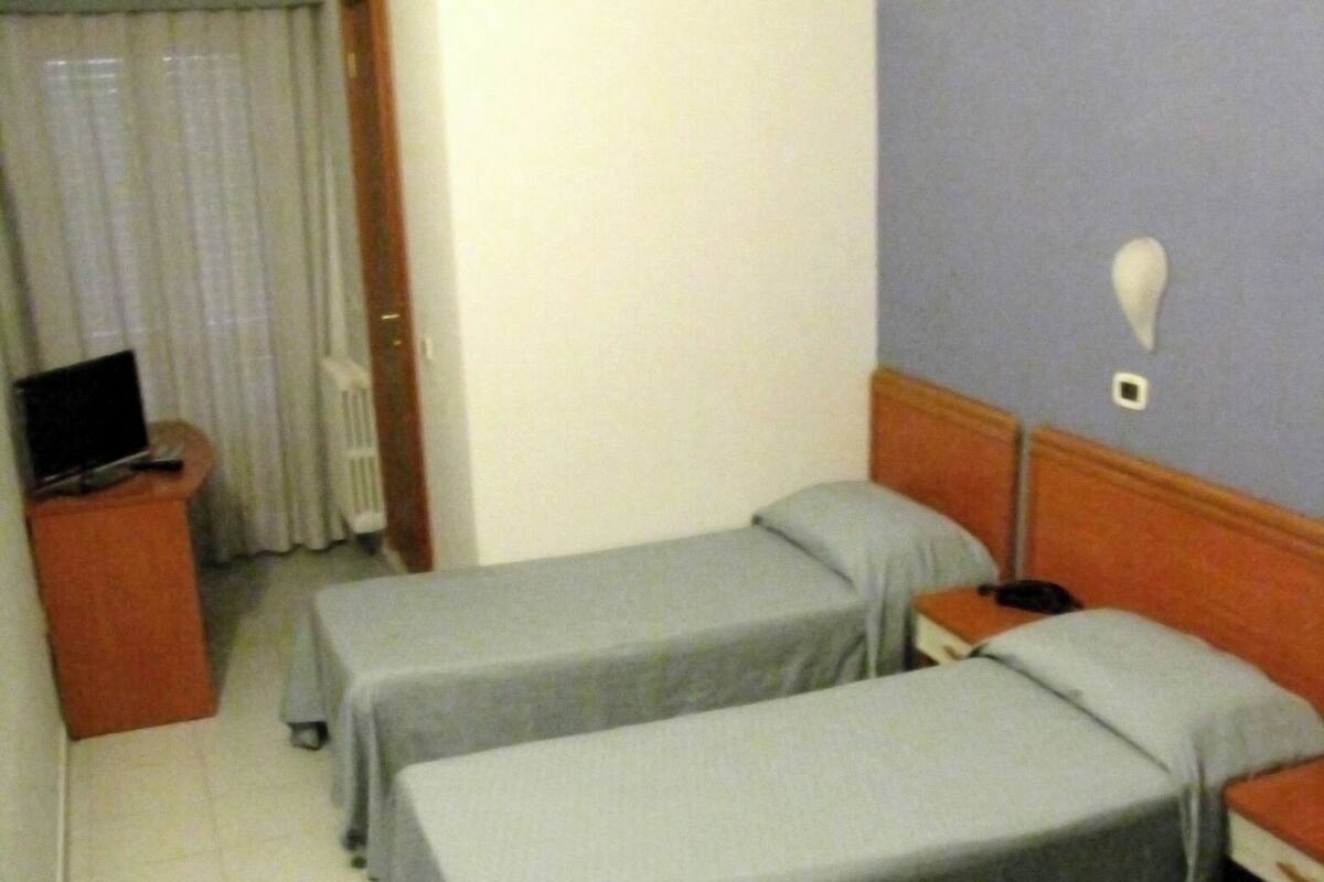 A hotel room with two single beds, a desk with a computer, a radiator, and a window with curtains.