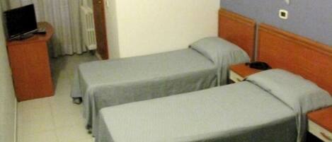 A hotel room with two single beds, a desk with a computer, a radiator, and a window with curtains.