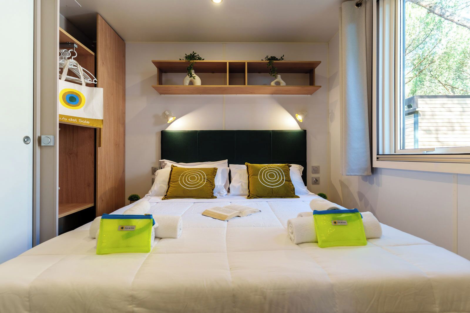 A neatly made bed with pillows, a bedside table with a book, and a shelf above the bed.