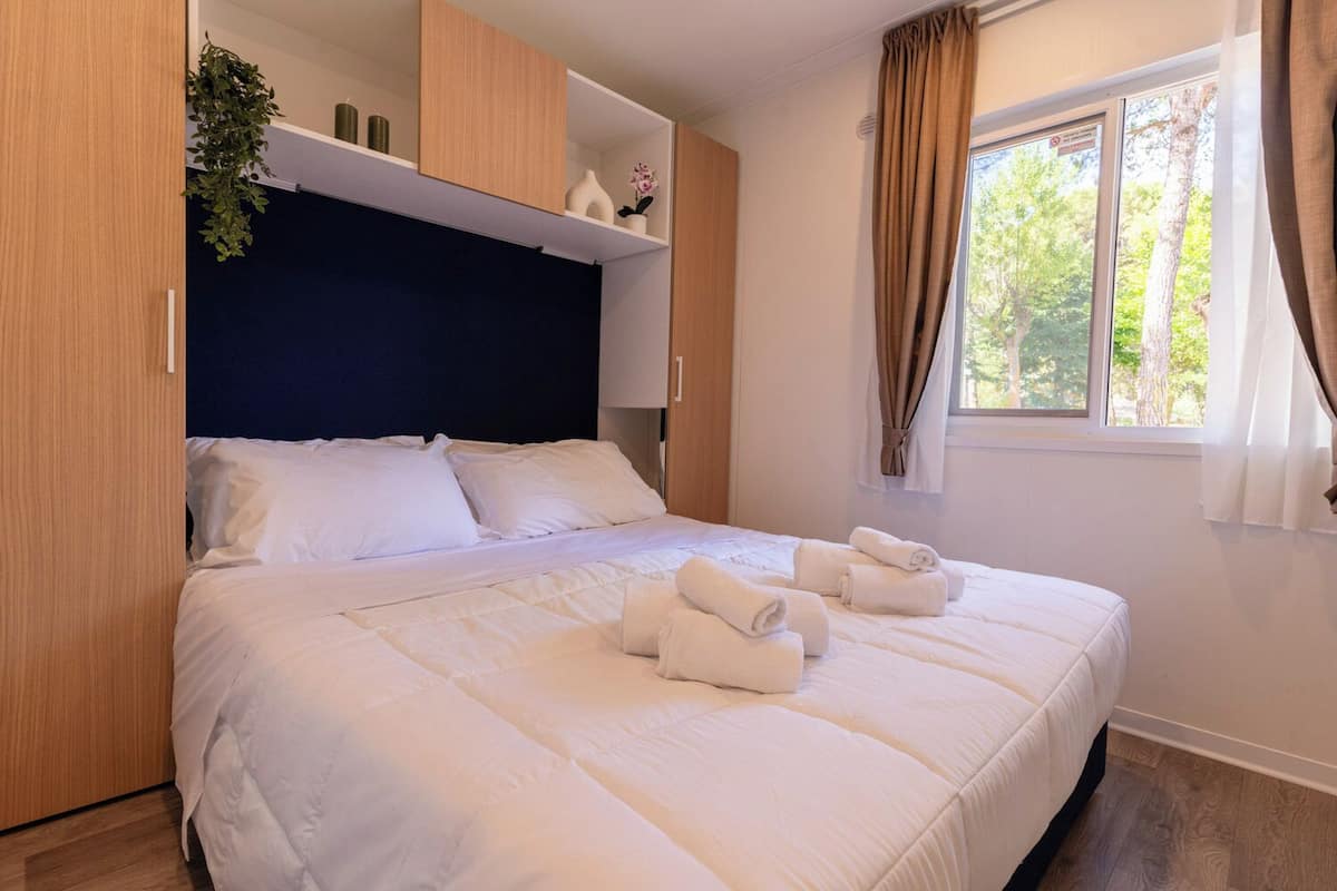 Una cama bien hecha con sábanas blancas y toallas dobladas, cabecera azul oscuro, armarios de madera y una ventana con cortinas.