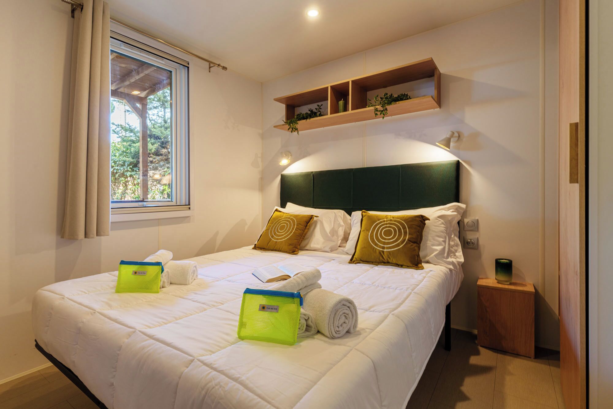 A neatly made bed with pillows, a bedside table, a shelf with plants, and a window with curtains.
