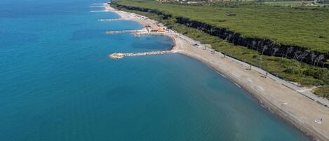 An aerial view of a coastal area with a sandy beach, a road parallel to the shore, and a green landscape with trees.
