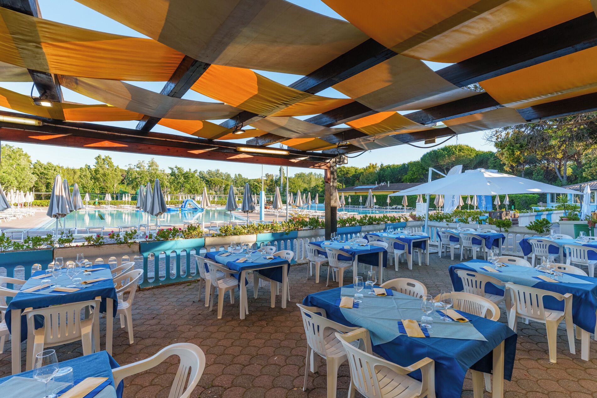 An outdoor dining area with tables and chairs, a pool in the background, and umbrellas providing shade.