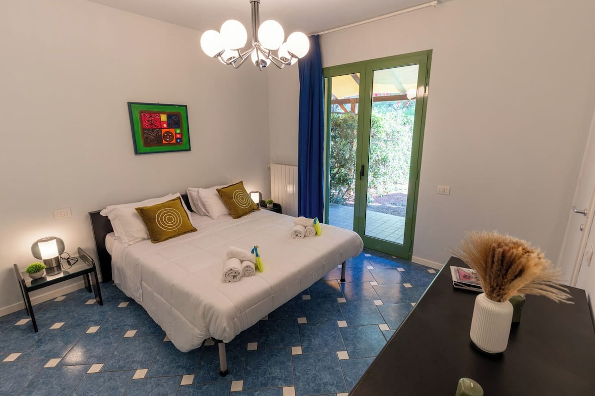 Habitación de hotel con cama, mesita de noche, una mesa con un florero y vistas al exterior.