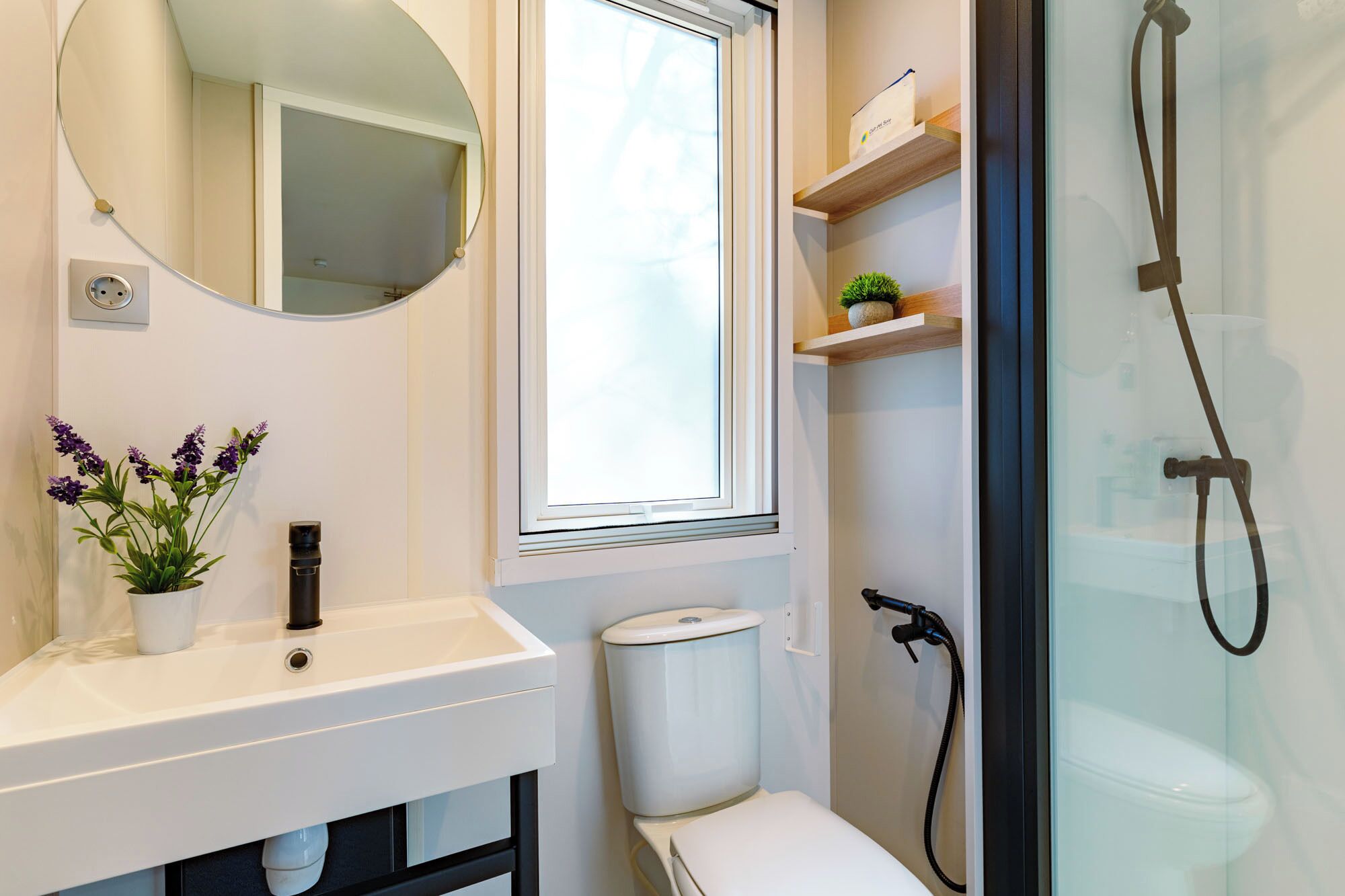 Ein modernes Badezimmer mit weißem Waschbecken, einem Spiegel, einer Toilette, einer Dusche und einem Fenster.