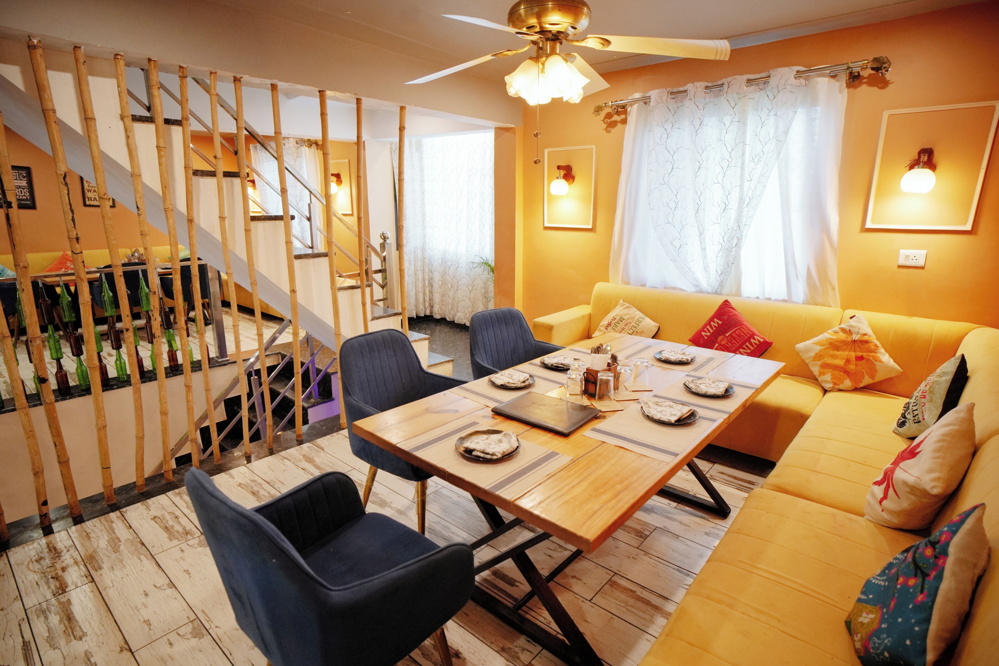 A dining area with a wooden table set for four, surrounded by chairs, and a sofa with cushions. A staircase with a bamboo railing is visible in the background.
