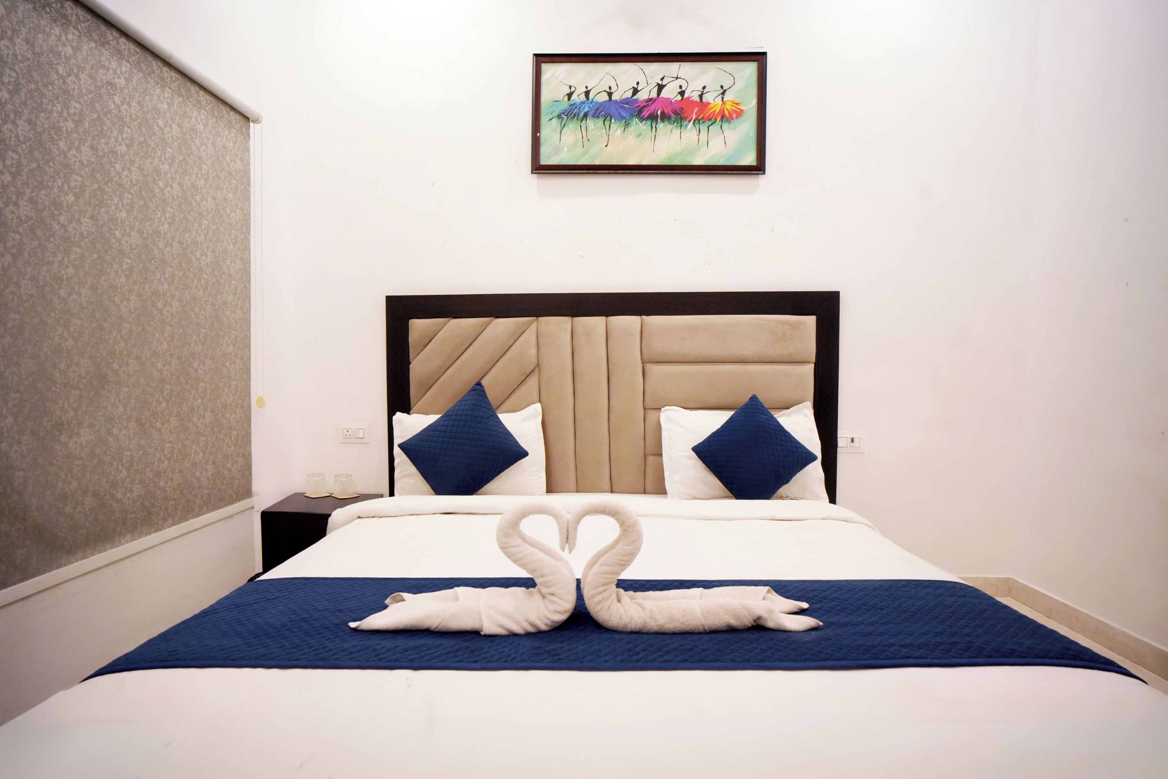 A hotel room with a large bed, a headboard, two pillows, a blue bedspread, and a wall-mounted painting.