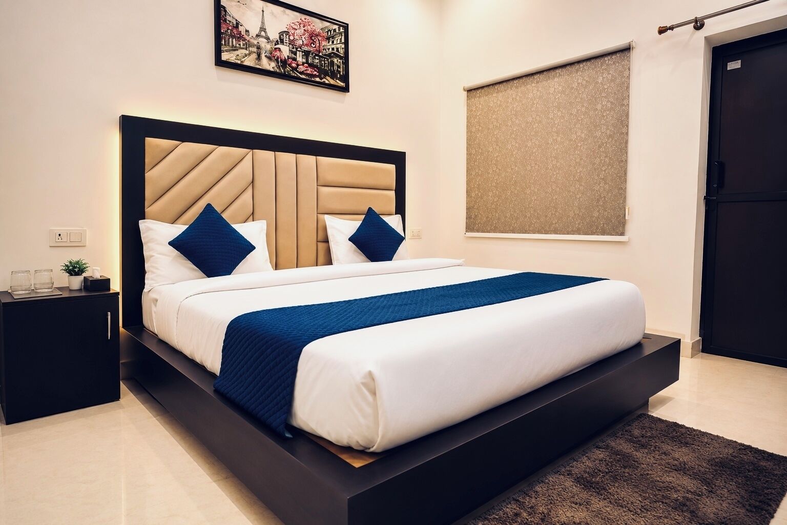 A modern hotel room with a large bed, a headboard with geometric patterns, a small bedside table with a plant, and a framed picture on the wall.