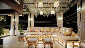 A cozy outdoor seating area with a sofa and wooden tables, surrounded by potted plants and hanging lights. - The Nines Hotel & Restro (Haldwani)