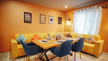 Un comedor con un sofá amarillo, sillas azules, una mesa de madera y un ventanal con cortinas.