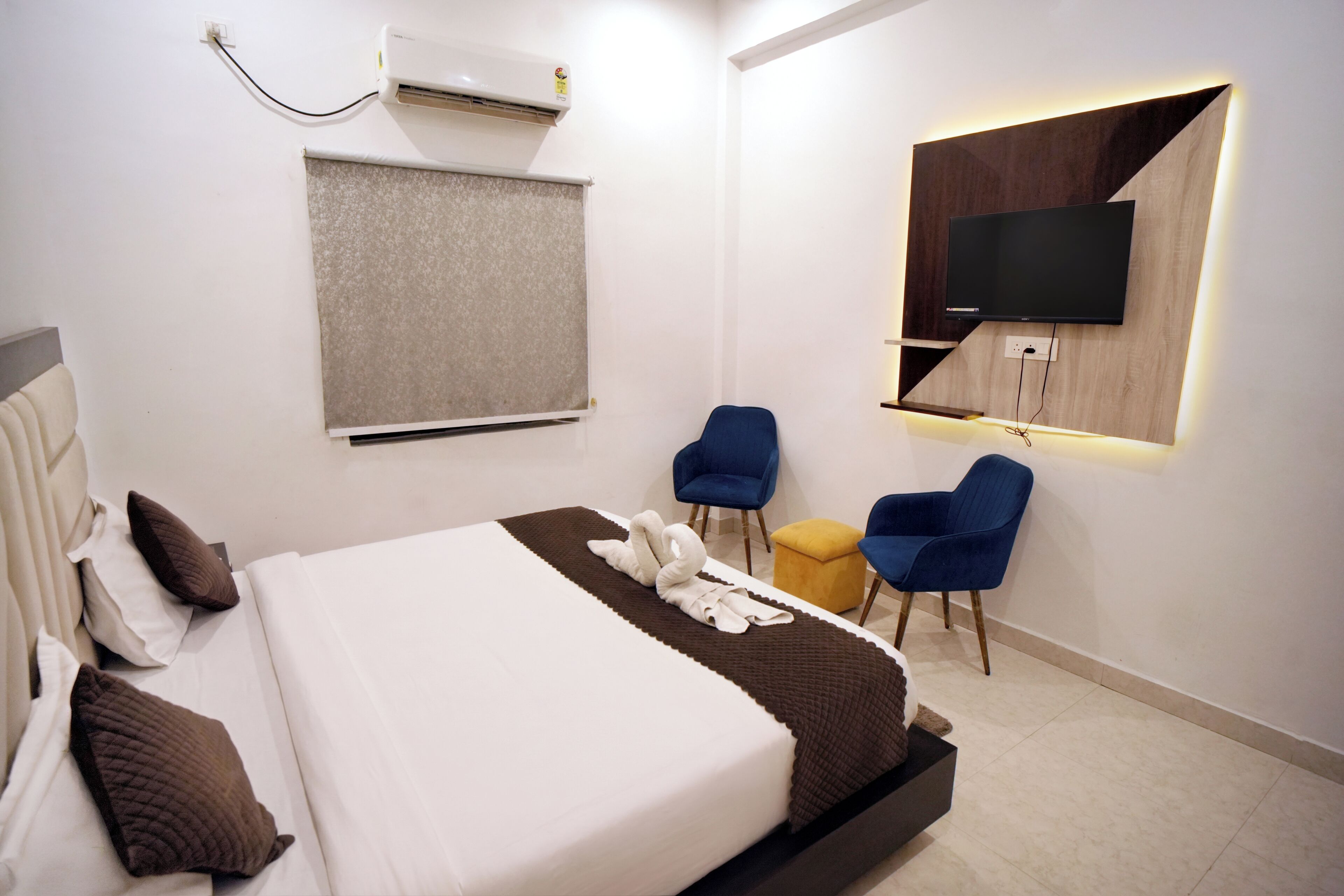 A hotel room with a bed, a flat-screen TV mounted on the wall, a desk with a chair, and a window with a blind.