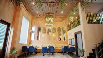 A restaurant interior with a long dining table, blue chairs, and a ceiling with hanging plants.