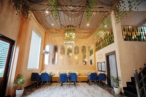 A restaurant interior with a long dining table, blue chairs, and a ceiling with hanging plants. - The Nines Hotel & Restro (Haldwani)