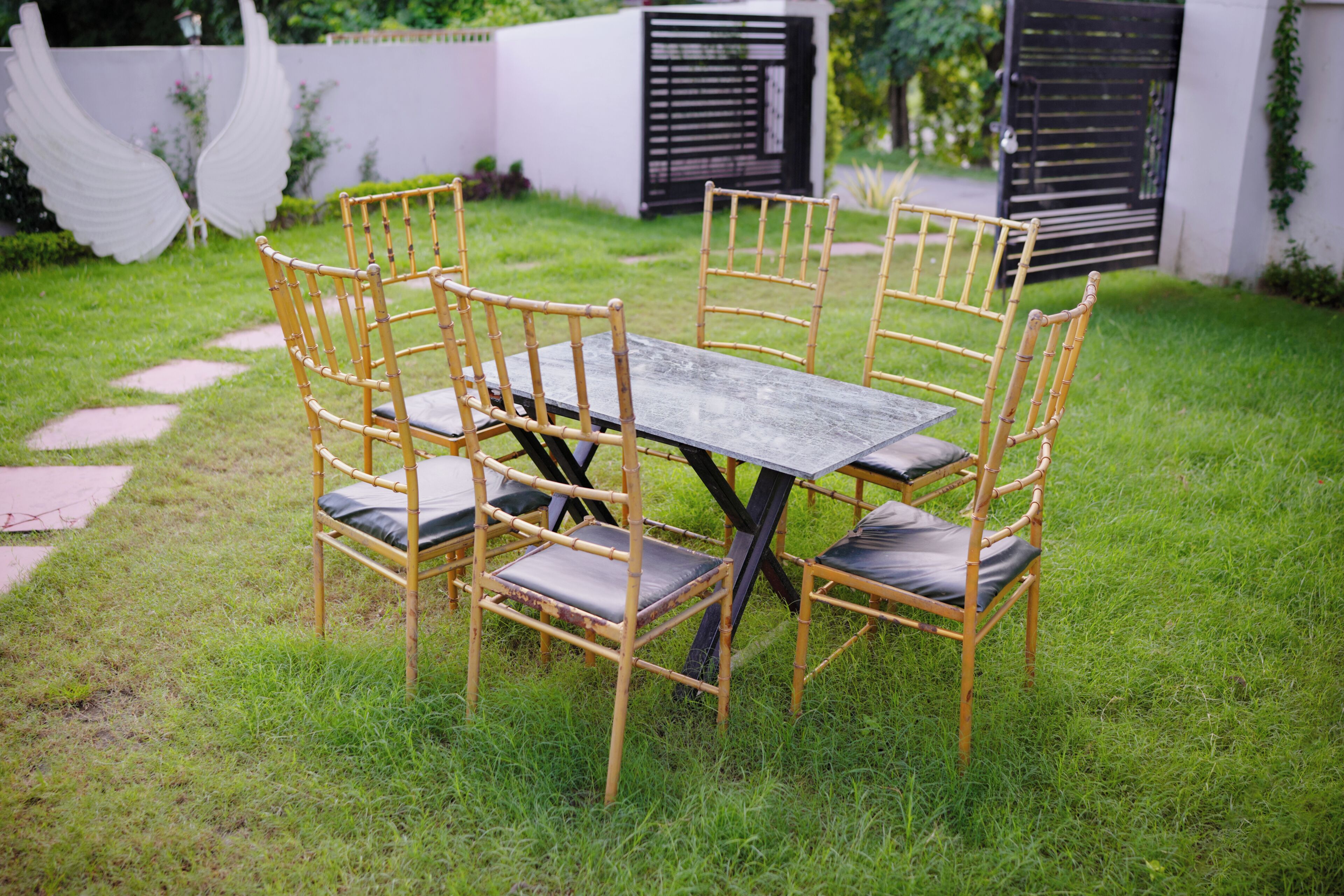 A rectangular outdoor table with four chairs, surrounded by a grassy area with a pathway leading to a gate.